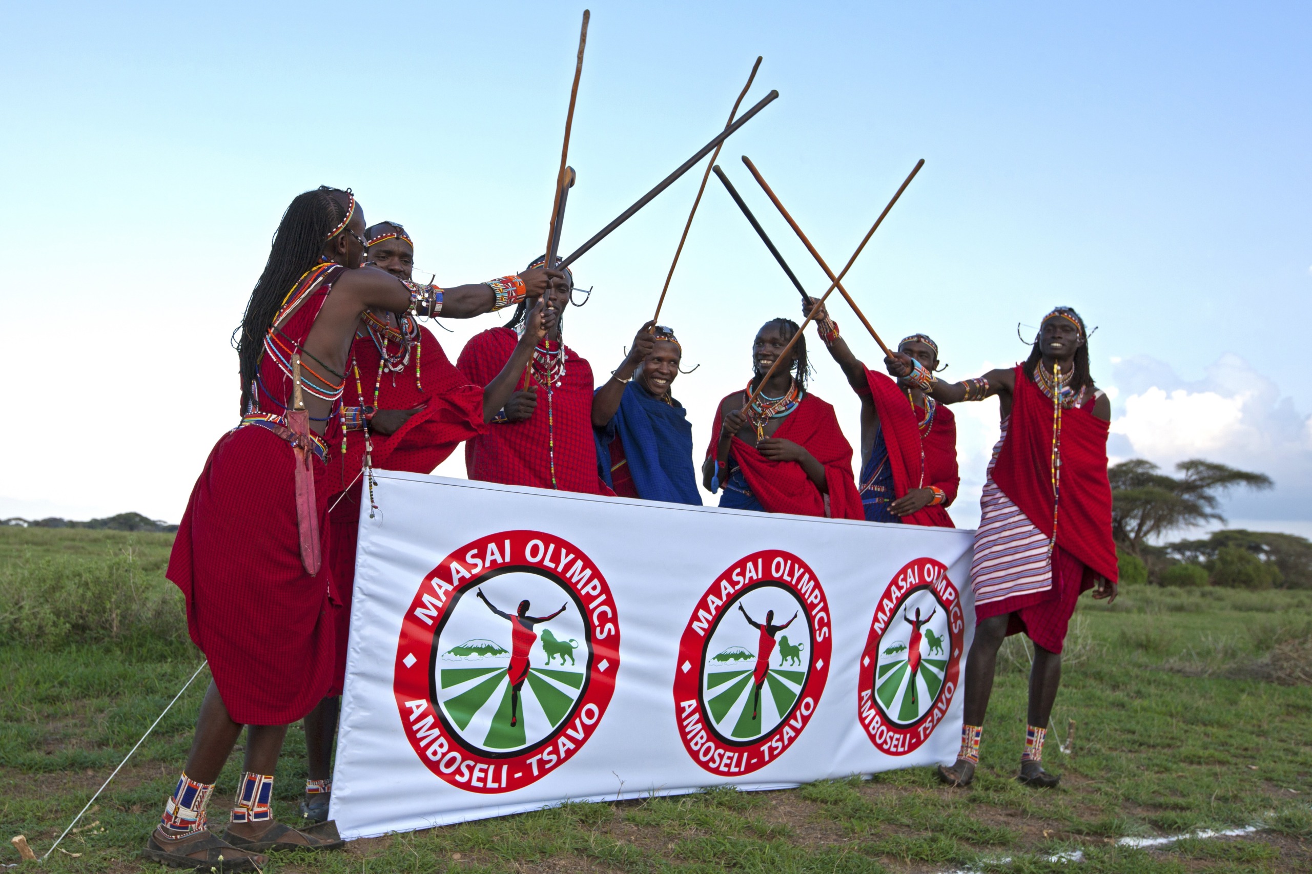 A group of Maasai people in traditional attire, with an air of pride, hold wooden sticks and gather around a Maasai Olympics Amboseli-Tsavo banner in an open field. This vibrant tribe appears to be celebrating or posing cheerfully.