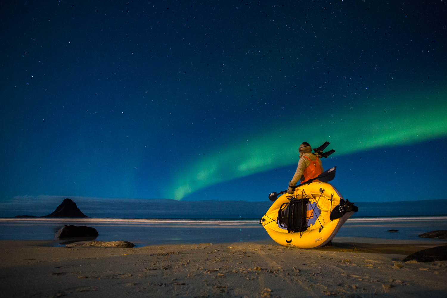 A person in a jacket and beanie sits in a yellow kayak on a sandy beach, gazing at the northern lights. The clear sky teems with stars, while the silhouette of a hill looms like an ancient whale imparted with the wisdom of ages in the background.