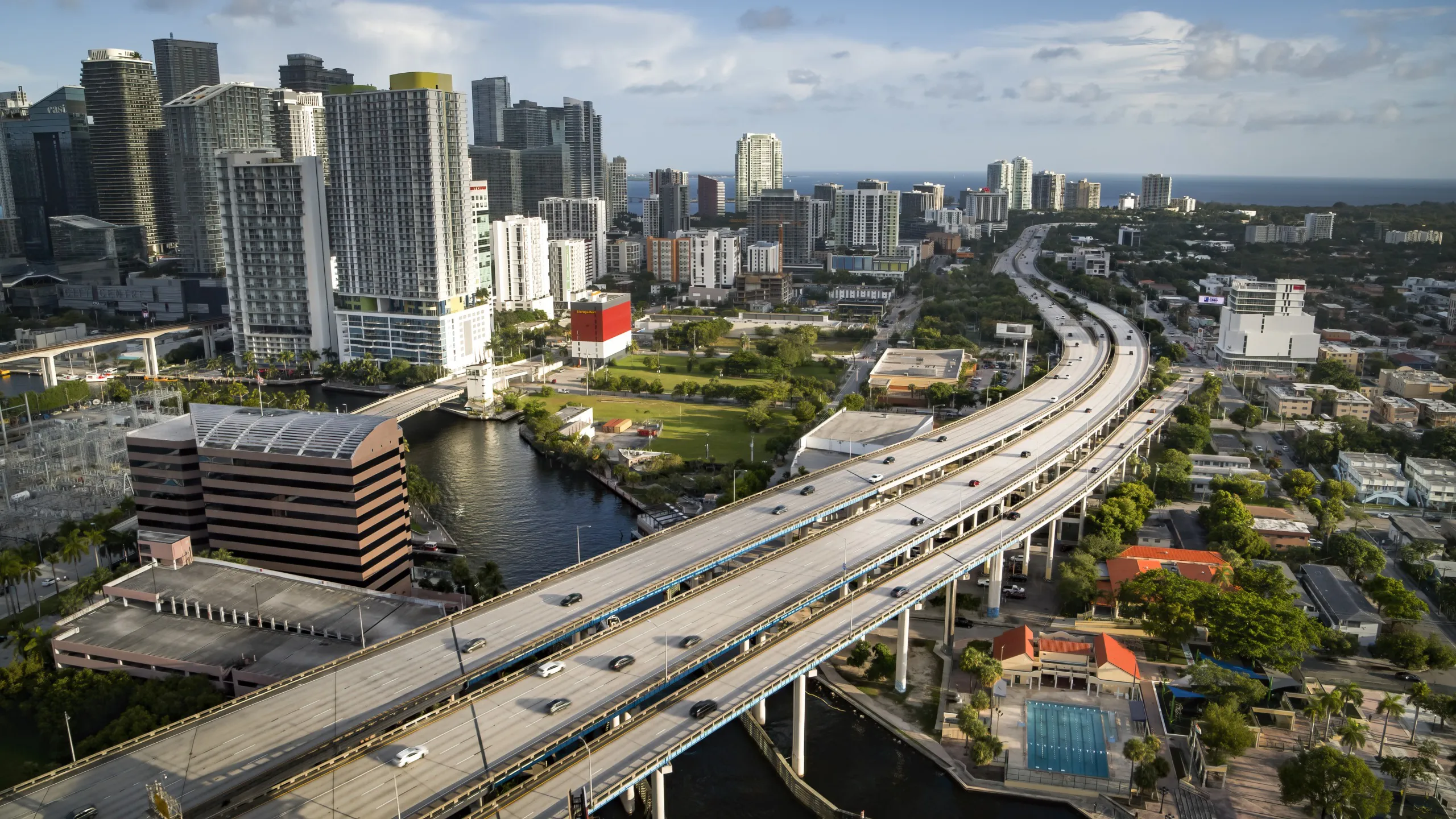 Aerial view of a vibrant cityscape reminiscent of Miami, with a highway weaving through towering skyscrapers and residential buildings. The scene unfolds with a river, green spaces, and a partially cloudy sky, while the wild beauty of the ocean stretches into the distance.