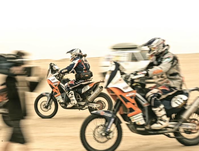 Two motorcyclists in racing gear speed across a desert landscape, reminiscent of the legendary Paris and Dakar. Dust swirls around as a blurred figure captures the action with a camera, while an off-road vehicle lingers in the background.