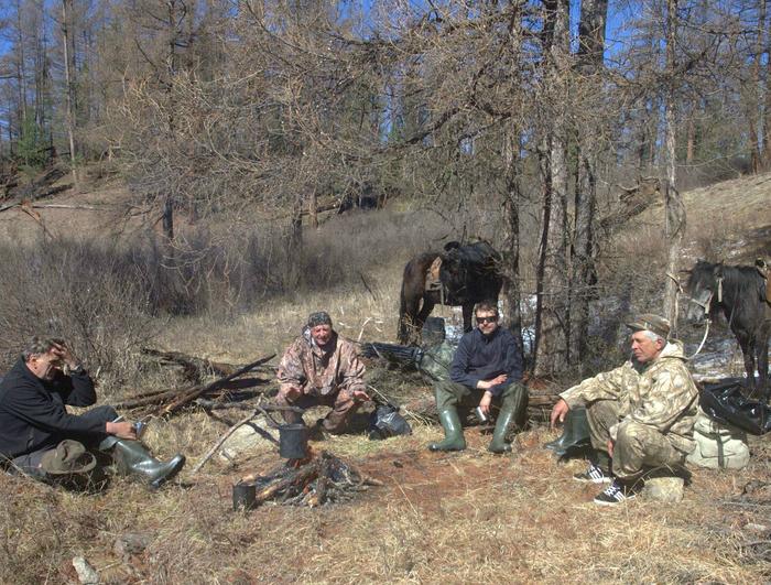 Four people, dressed in outdoor gear with some sporting hats and boots, sit around a campfire in the heart of Asia's Amazon. Nearby, two horses stand calmly among the trees and grass, as if waiting for black-billed capercaillies to join the scene.