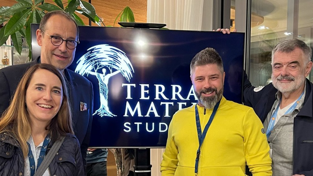 Four people stand indoors before a screen displaying the Terra Mater Studios logo. Smiling, they seem to be at an event celebrating nature and science. Behind them, greenery complements the sleek modern building interior.