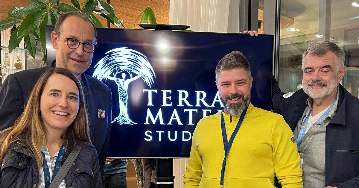 Four people stand indoors before a screen displaying the Terra Mater Studios logo. Smiling, they seem to be at an event celebrating nature and science. Behind them, greenery complements the sleek modern building interior.