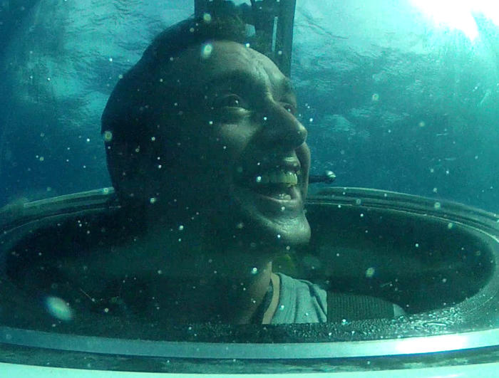 A person smiling inside a small submersible vessel, surrounded by water. The image captures their face through a clear, round window as sunlight filters through the water, creating a serene, Terra Mater-like underwater atmosphere reminiscent of nature films.