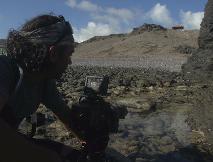 A person with a bandana is crouched beside a professional camera at a rocky Brazilian shore, capturing the rugged terrain and tide pools under cloudy skies.