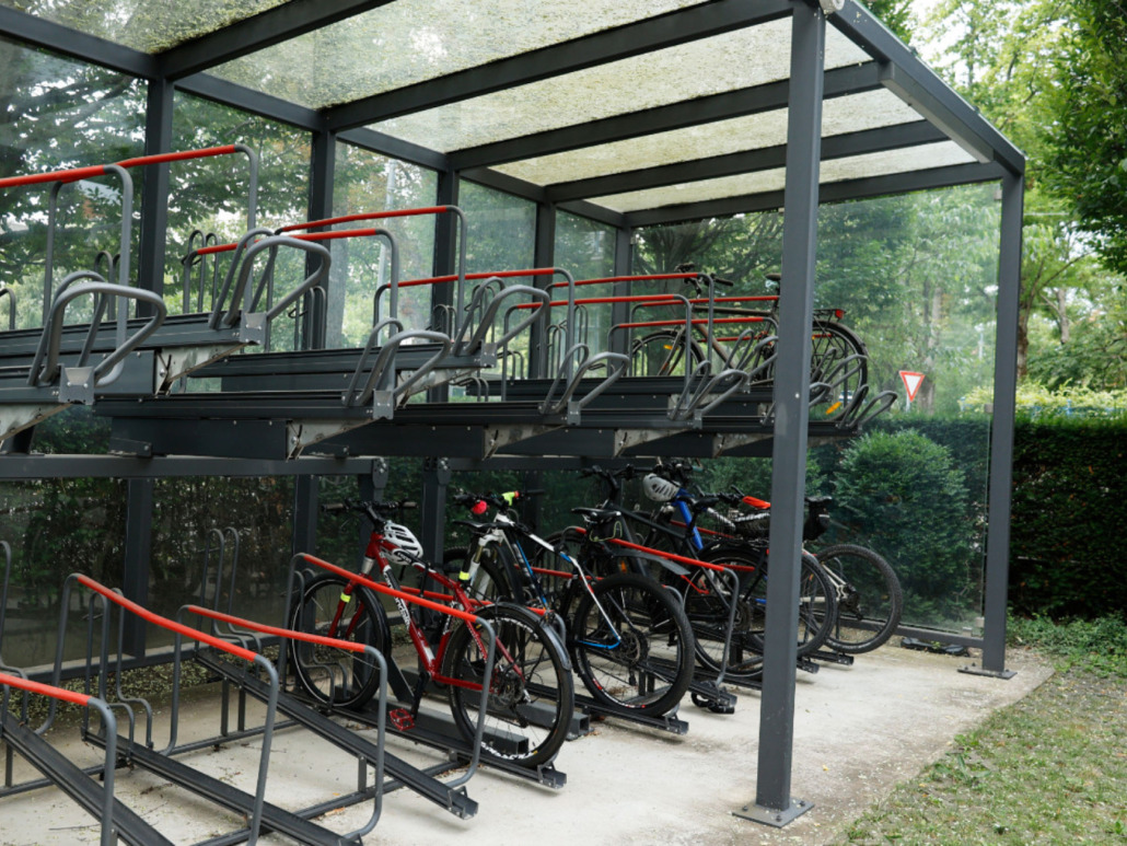 A two-tiered bike rack shelter with several bicycles parked on the bottom level. The shelter has a clear roof, surrounded by trees and greenery.