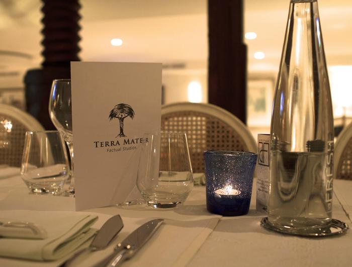 In a softly lit restaurant, a table set for dining showcases a menu labeled Terra Mater Factual Studios, alongside reviewed selections from MIPTV 2013. The arrangement features a blue candle holder with a glowing candle, elegant glassware, cutlery, and a water bottle on a pristine white tablecloth.