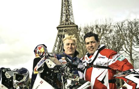 Two motorcyclists in racing gear stand next to their bikes in front of the Eiffel Tower, exuding reel feelings as they smile for a factual entertainment series. The sky is overcast and trees without leaves are visible in the background, setting the scene for this lively event.