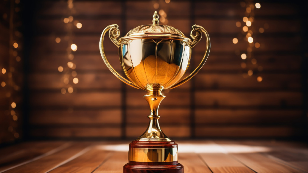 A shiny gold trophy with ornate handles sits on a wooden table, gleaming like the ultimate prize. The background is softly blurred with warm glowing lights and wooden paneling, creating a celebratory and elegant ambiance reminiscent of grand festival news.