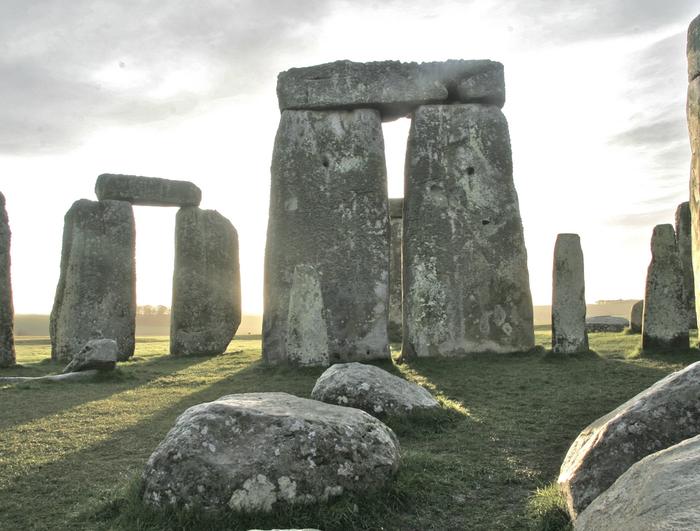 As the sun sets over Stonehenge, the large standing stones create a mesmerizing silhouette in a circular formation on the grassy field. The partly cloudy sky enhances the dramatic scene, casting long shadows that seem to mysteriously bring back echoes of the past.