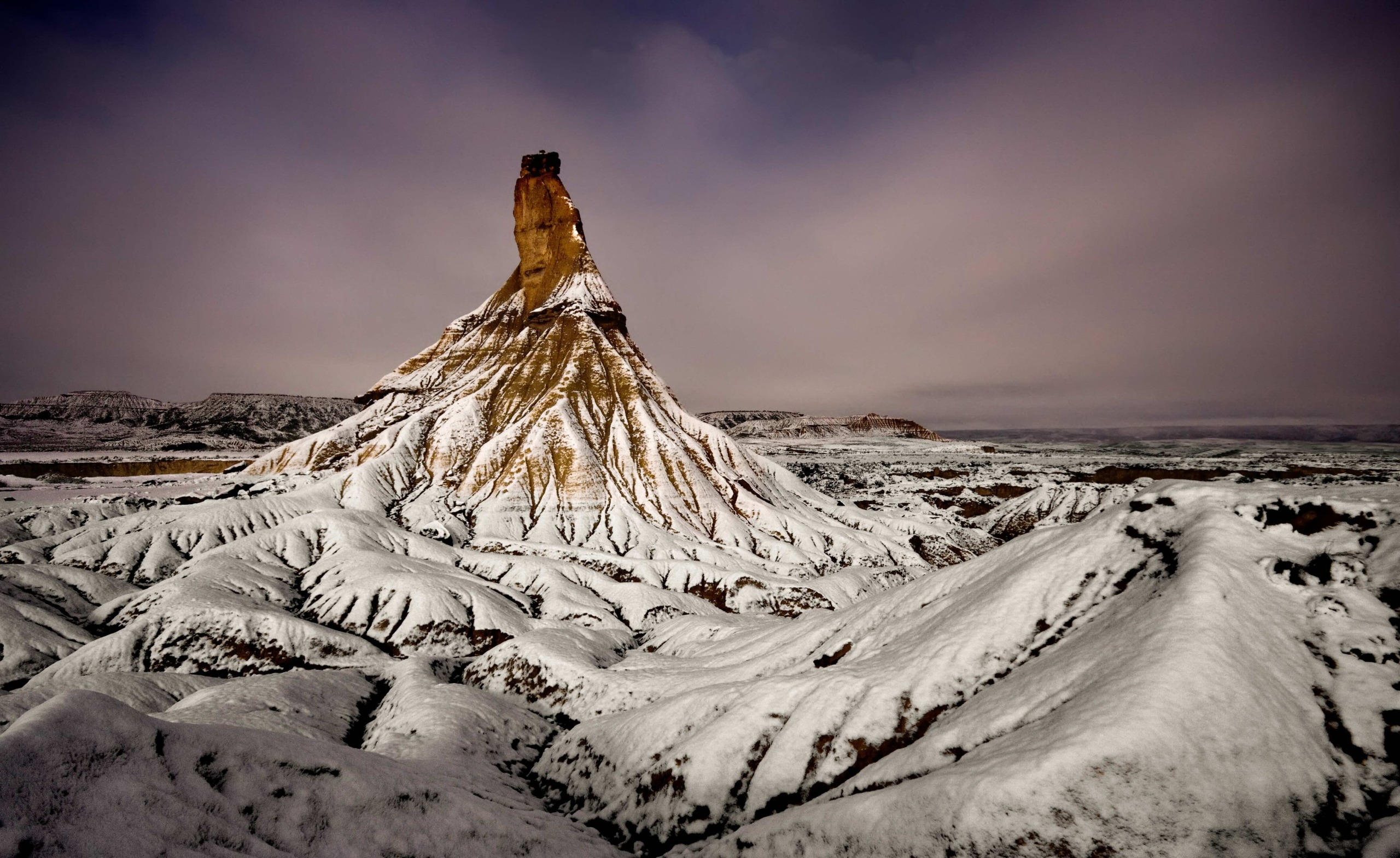 A dramatic landscape featuring a striking rock formation in Europe's Badlands, surrounded by snow-covered, rugged terrain. The tower-like structure stands prominently against a moody, overcast sky, creating a stark and captivating contrast.