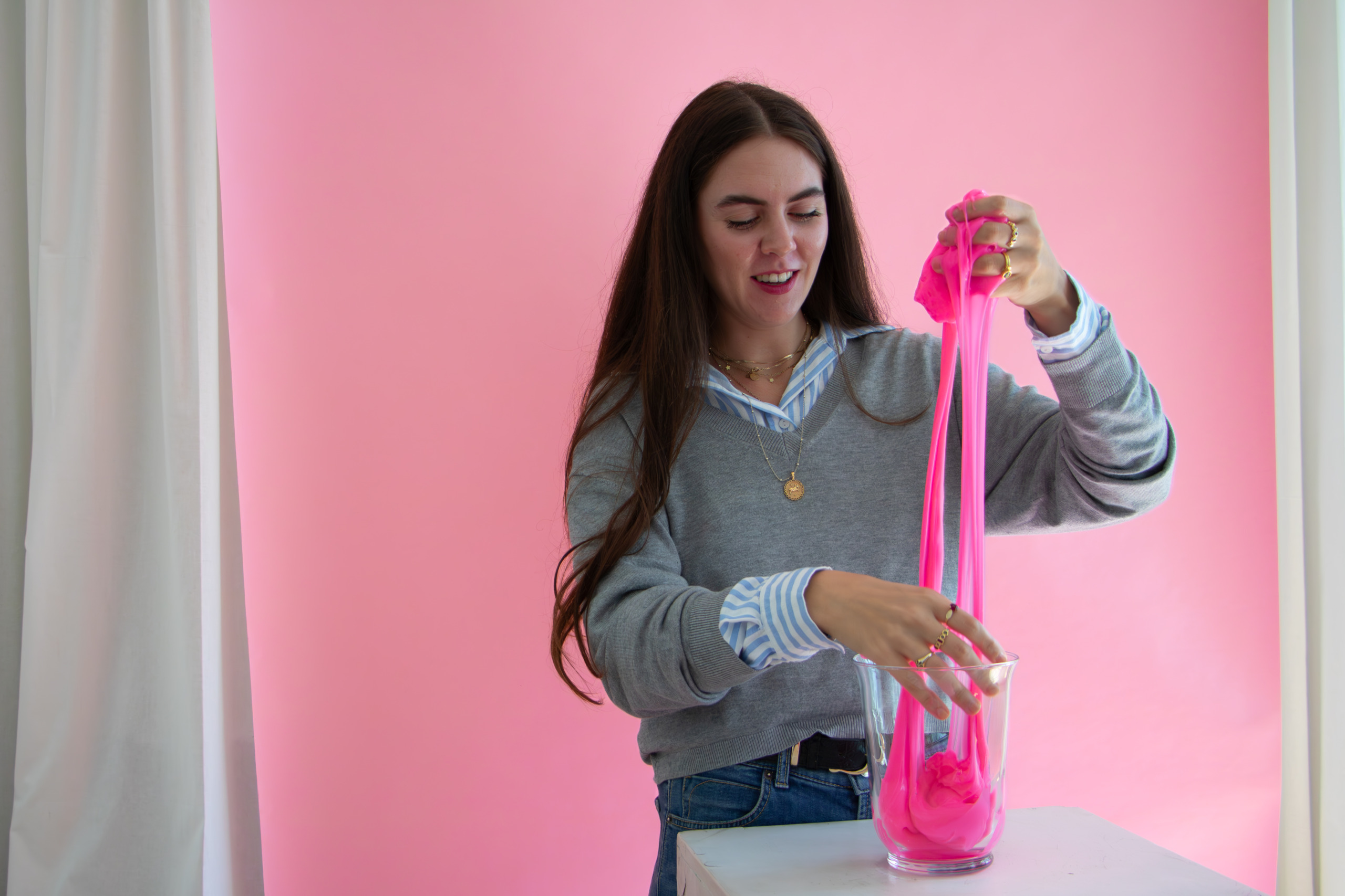 A person with long dark hair smiles while stretching bright pink slime over a glass container. They wear a gray sweater over a striped shirt. The background is a soft pink, creating a playful and colorful scene.