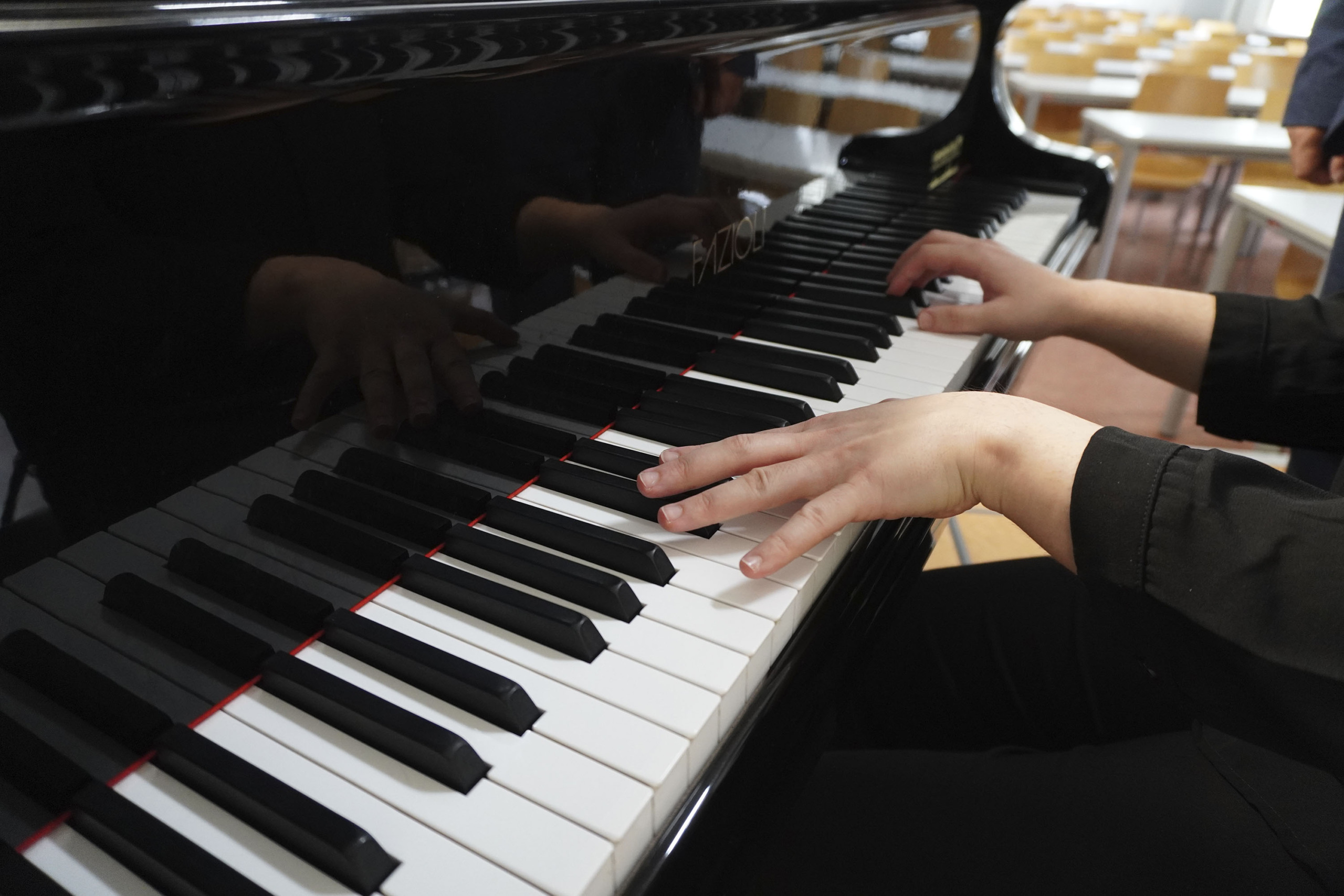 A person wearing a dark shirt plays a black piano with white and black keys. Their hands are positioned over the keyboard in a well-lit room with wooden chairs and tables in the background.