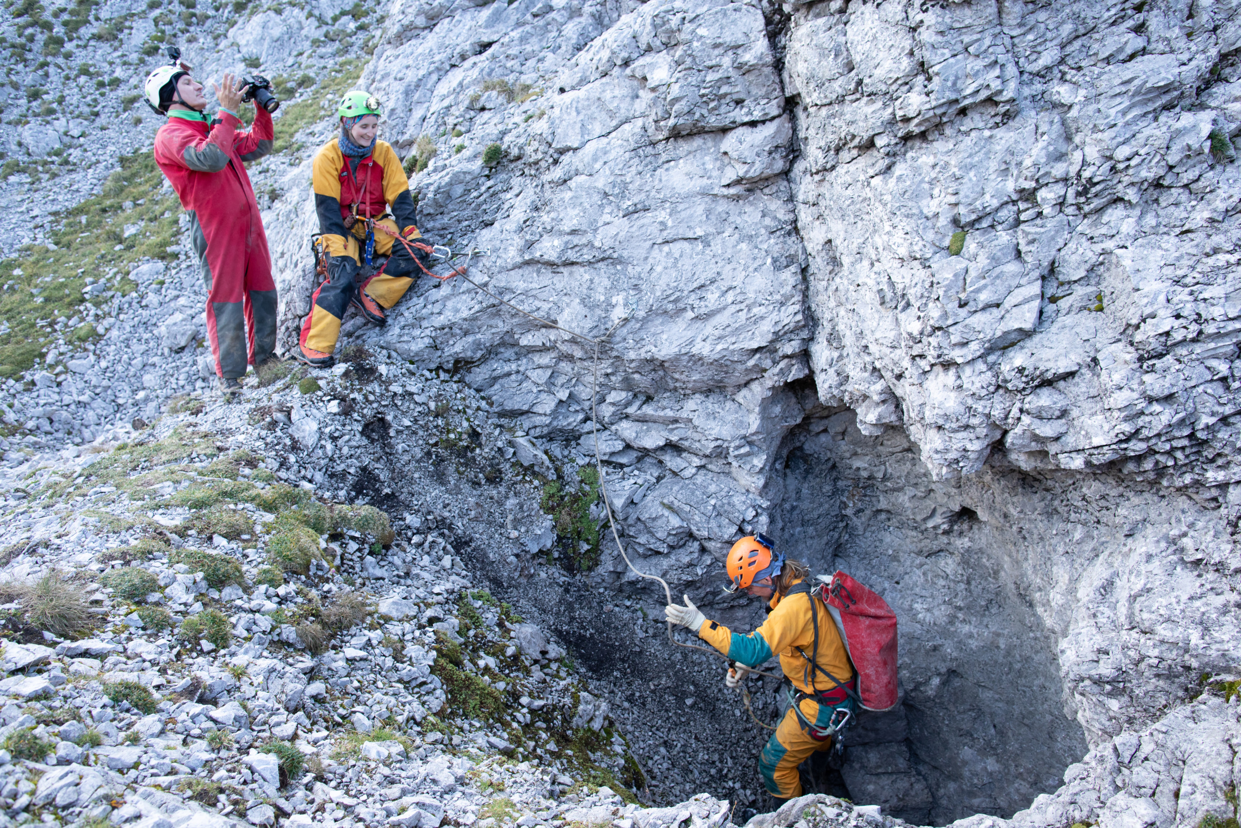 Three cavers, wearing helmets and protective suits, explore a rocky terrain. One person marks a position near a cave entrance while another takes photographs. They are surrounded by rugged stone formations.