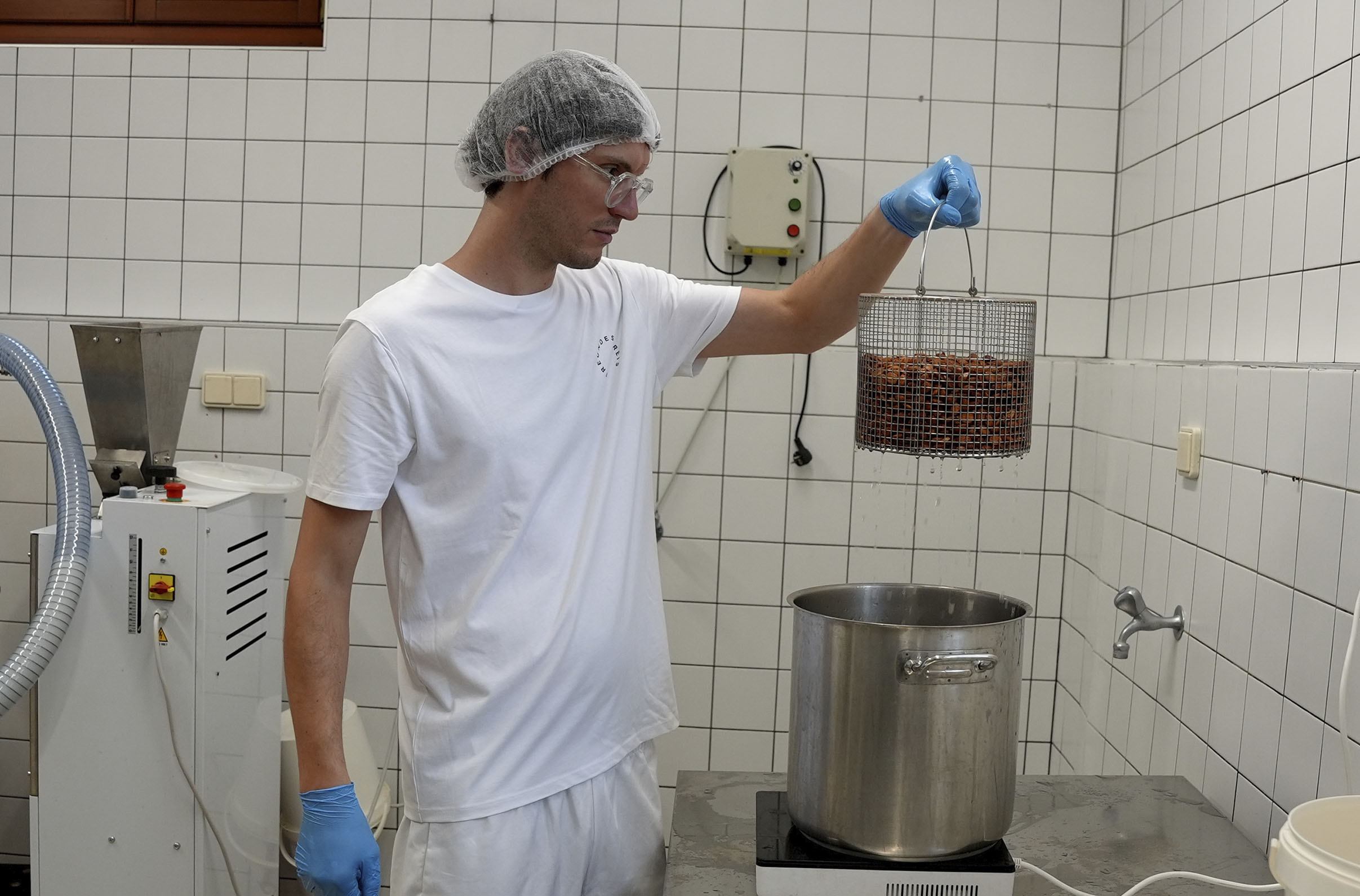 A person wearing a white uniform, hairnet, gloves, and glasses holds a basket of nuts over a large pot in a tiled kitchen. A machine and an electrical panel are visible in the background.