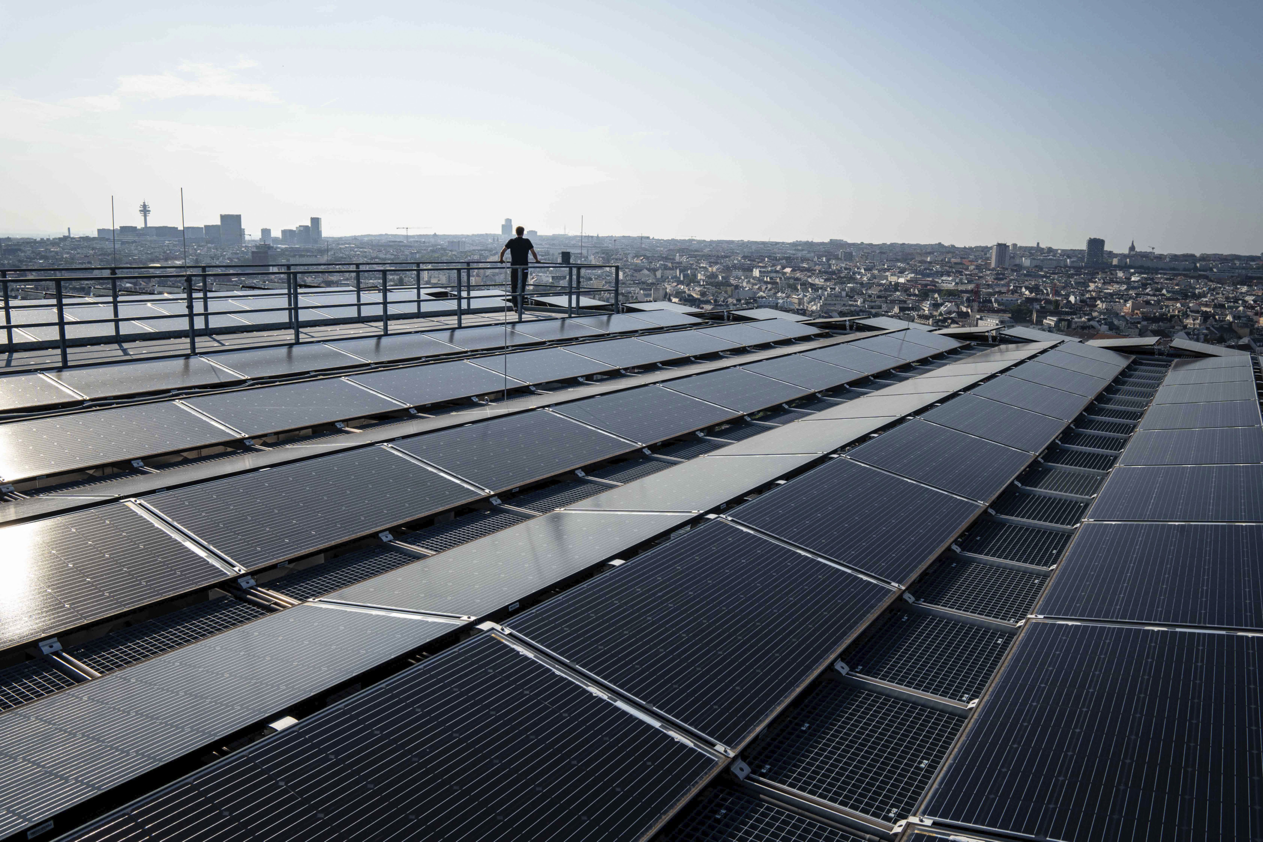 A person stands on a rooftop overlooking a city, surrounded by rows of solar panels under a clear sky. The skyline features various buildings and a distant tower.