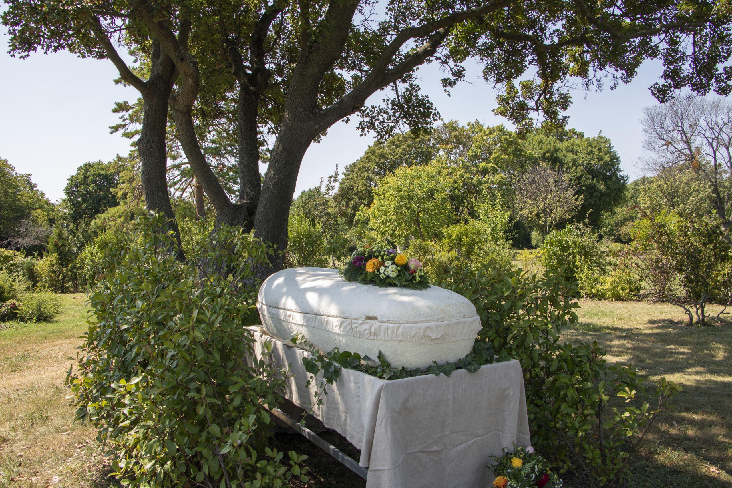 A white coffin adorned with colorful flowers is set outdoors under a large tree, surrounded by lush greenery, on a sunny day. A white cloth drapes the platform supporting the coffin.