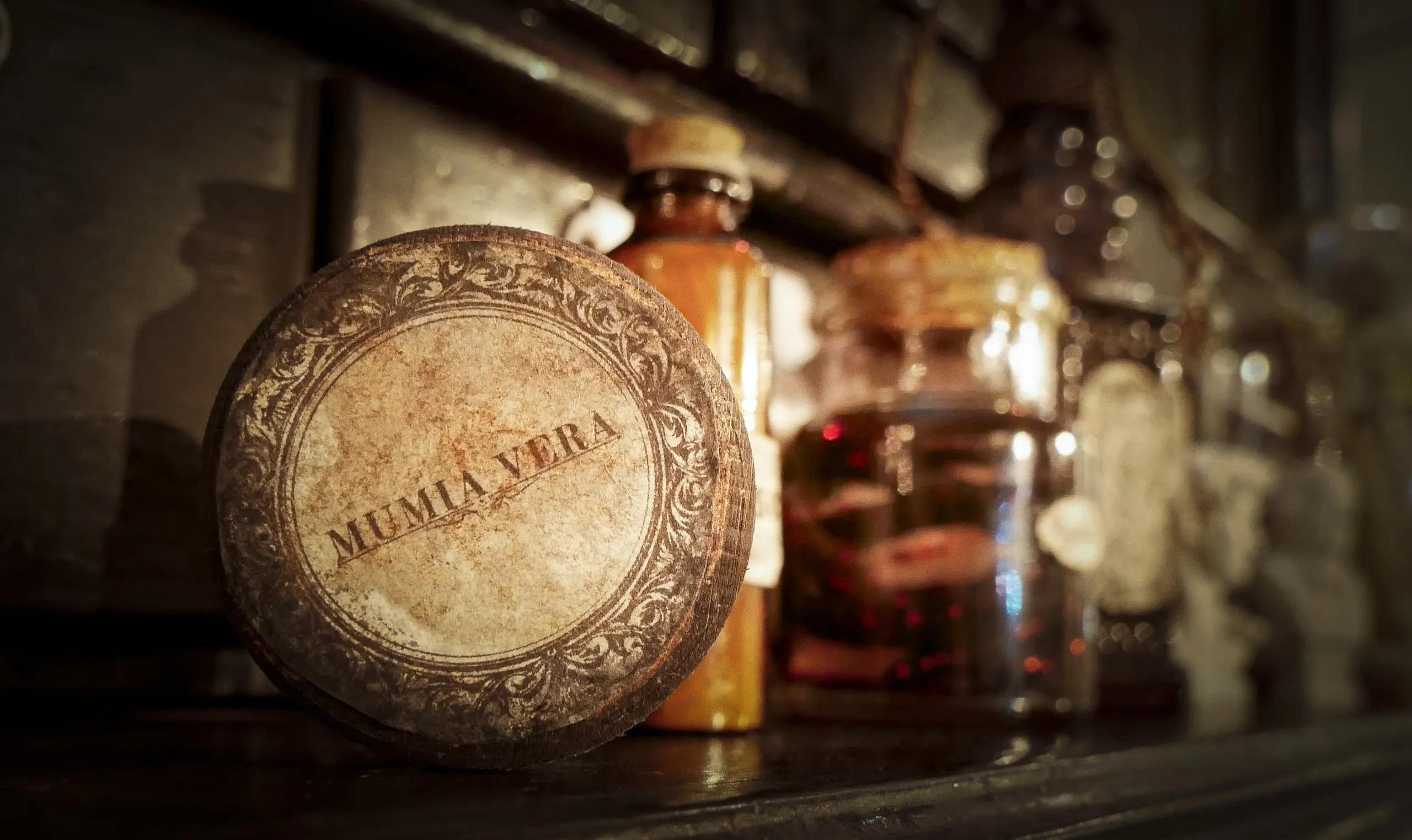 A collection of vintage apothecary bottles and containers, including a round container labeled Mumia Vera, sit on a dark wooden shelf. The labels and glass jars have an antique appearance, evoking an old-world or historical setting.