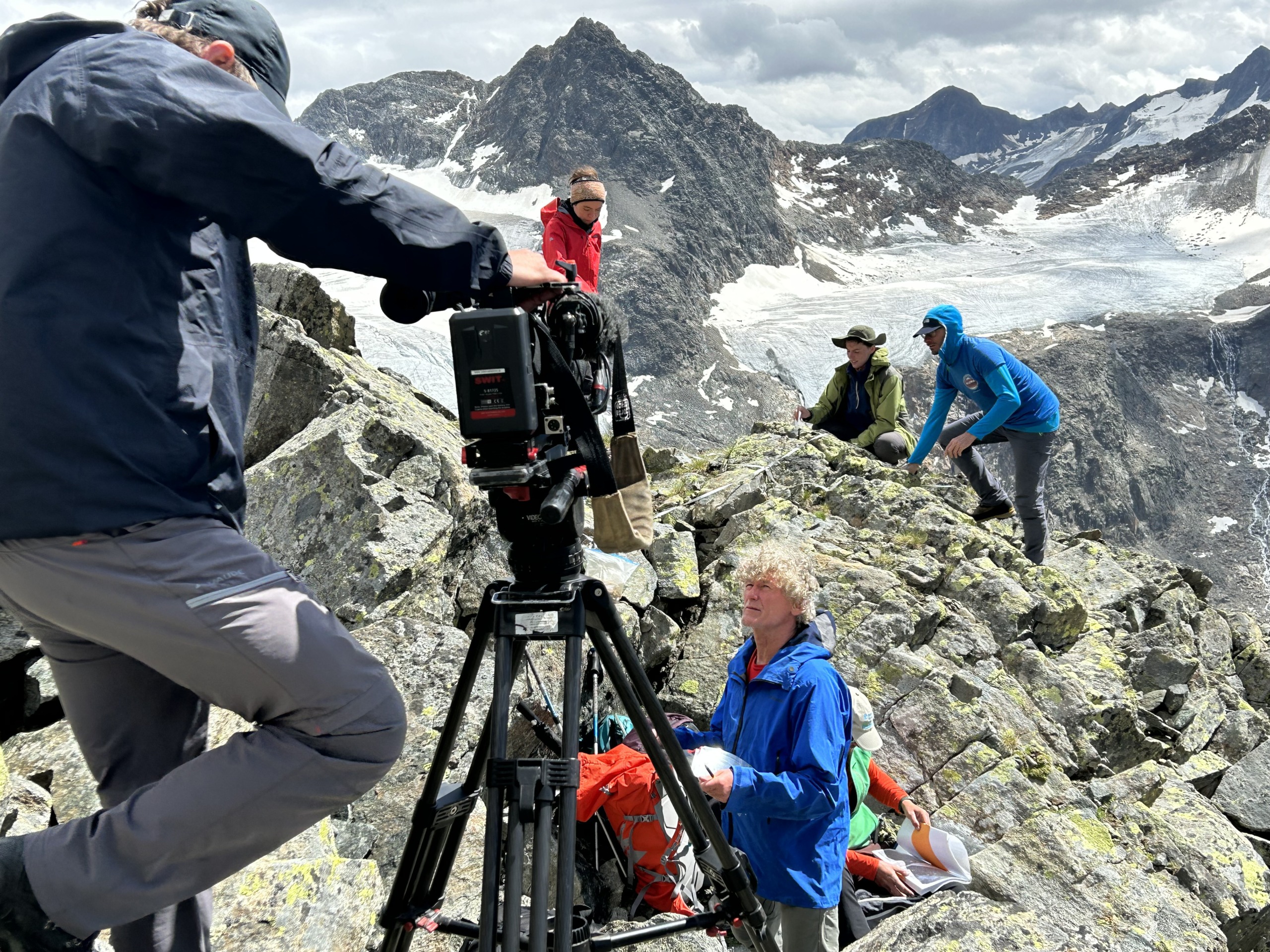 A dedicated film crew is setting up equipment on a rocky mountain peak, surrounded by majestic snow-capped mountains. The sky is partly cloudy as five people work together, with one expertly operating the camera and others assisting. They're committed to green producing throughout the shoot.