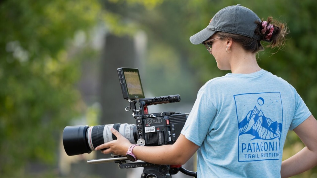 A person wearing a cap and a Patagonia Trail Running T-shirt operates a professional video camera with a large lens outdoors. Surrounded by greenery, they focus intently on the camera's monitor, capturing the essence of sustainability in each frame.