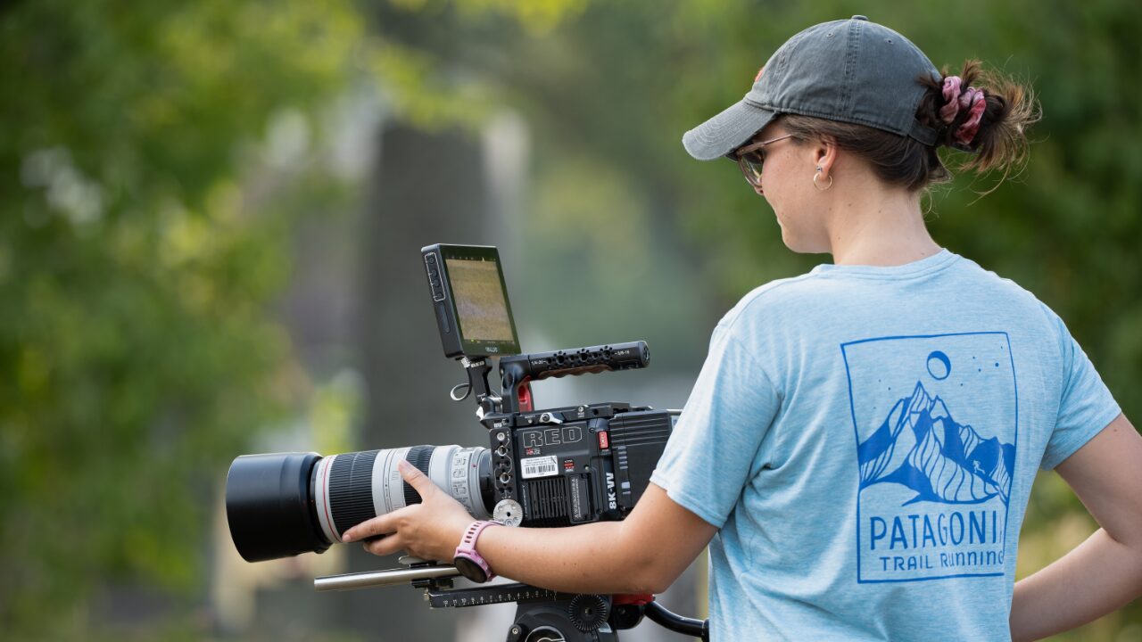 A person wearing a cap and a Patagonia Trail Running T-shirt operates a professional video camera with a large lens outdoors. Surrounded by greenery, they focus intently on the camera's monitor, capturing the essence of sustainability in each frame.