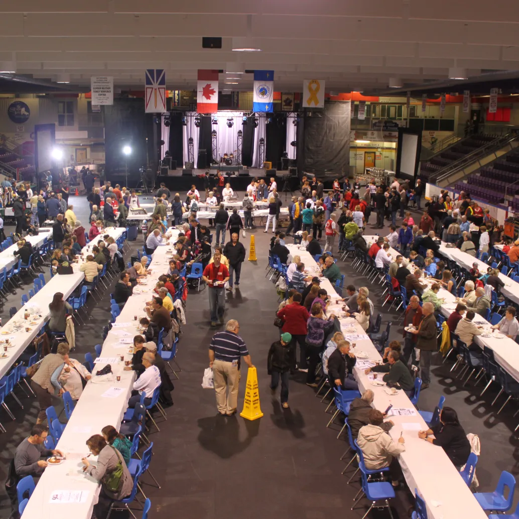 A large indoor event buzzes with activity as long tables and chairs fill with people. Various national flags hang from the ceiling, reminiscent of unity in challenging times like 9/11. A stage stands at one end, ready for unexpected visitors to share their stories amid this lively atmosphere.