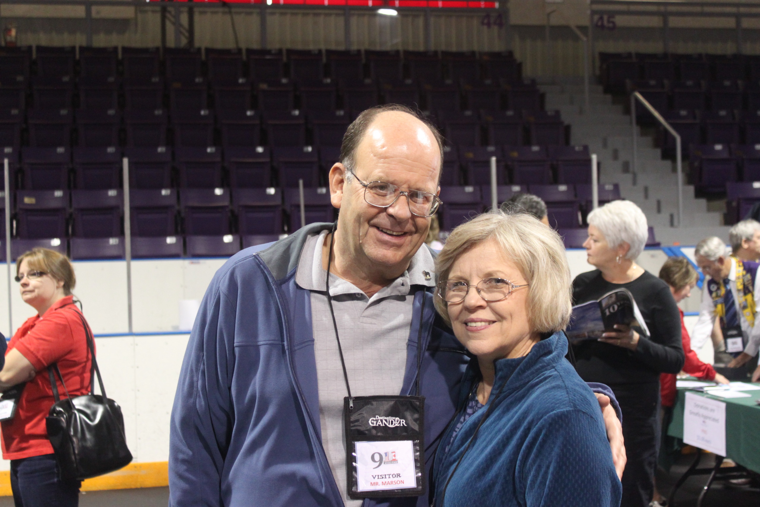 A man and a woman are smiling and standing close together in an indoor stadium, embodying the spirit of 9/11 Silver Linings. The man dons a gray shirt with a blue jacket, while the woman is clad in her own blue jacket. People gather in the background, amid empty seats hinting at unexpected visitors.