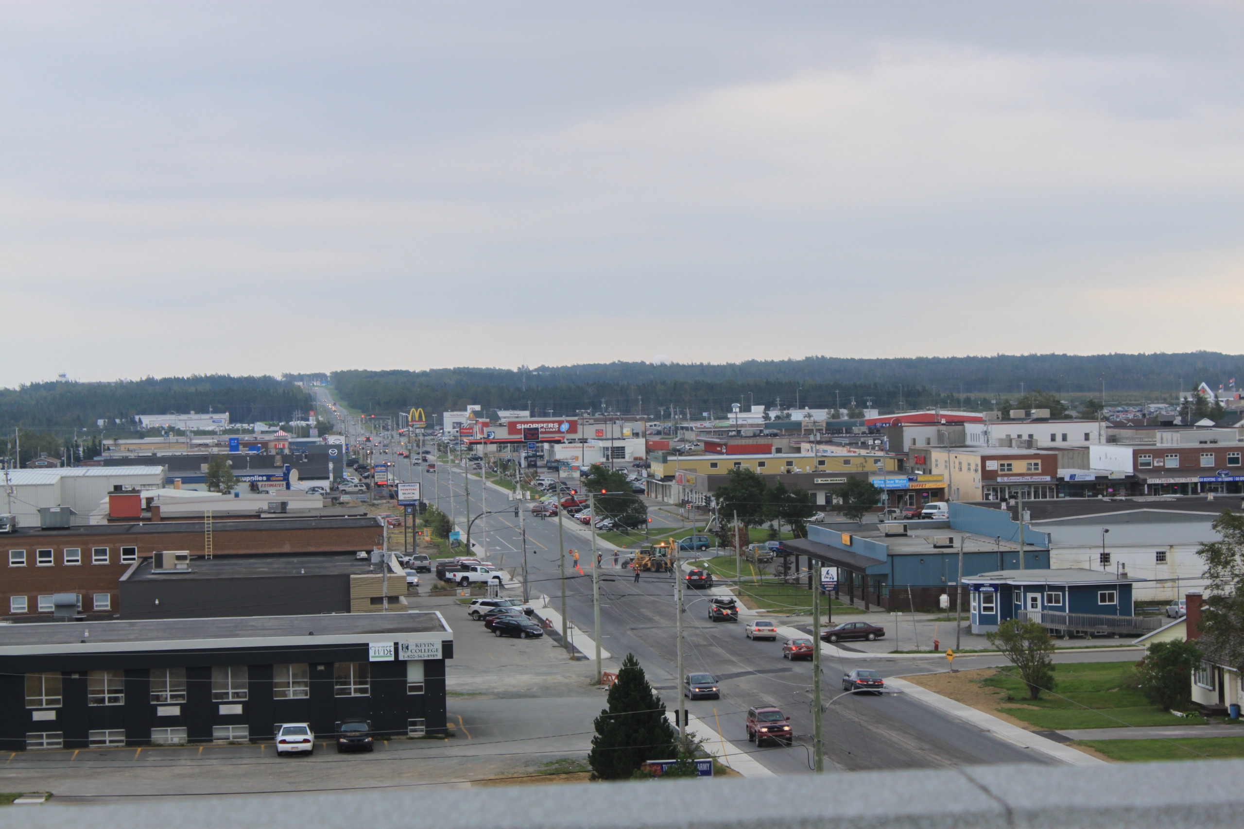 A view of a small town with a main street lined with buildings, including shops and offices. Cars are parked along the road, and the skyline is overcast with gray clouds. Yet, like silver linings after a storm, green trees offer a hint of serenity in the distance.