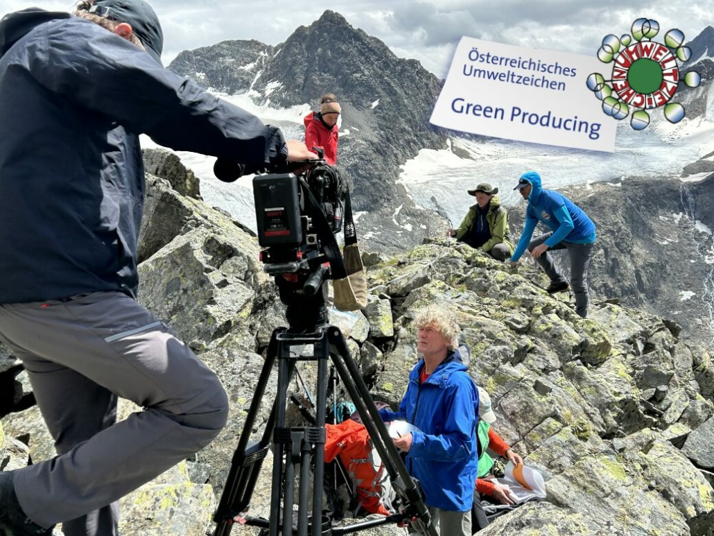 Amidst a rocky mountain landscape, a film crew is capturing a climber in a blue jacket. A sign proudly displays Österreichisches Umweltzeichen and highlights their commitment to green producing, as snow-capped peaks rise majestically in the background.