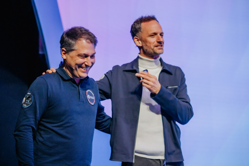 Ian Carnelli from ESA and Markus Mooslechner from Terra Mater Studios are standing on stage against a blue and purple backdrop at the Microsoft AI Tour. The person on the left is smiling in a dark sweater with a patch, while the person on the right has their arm around them, gesturing enthusiastically.