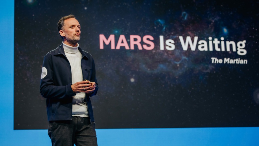 Markus Mooslechner stands on a stage in front of a large screen displaying the quote "MARS Is Waiting" from the book The Martian. Dressed in a white turtleneck and dark jacket, they hold a small device. The backdrop features a starry space image, hinting at the future of space exploration as part of the Hera Space Companion initiative.