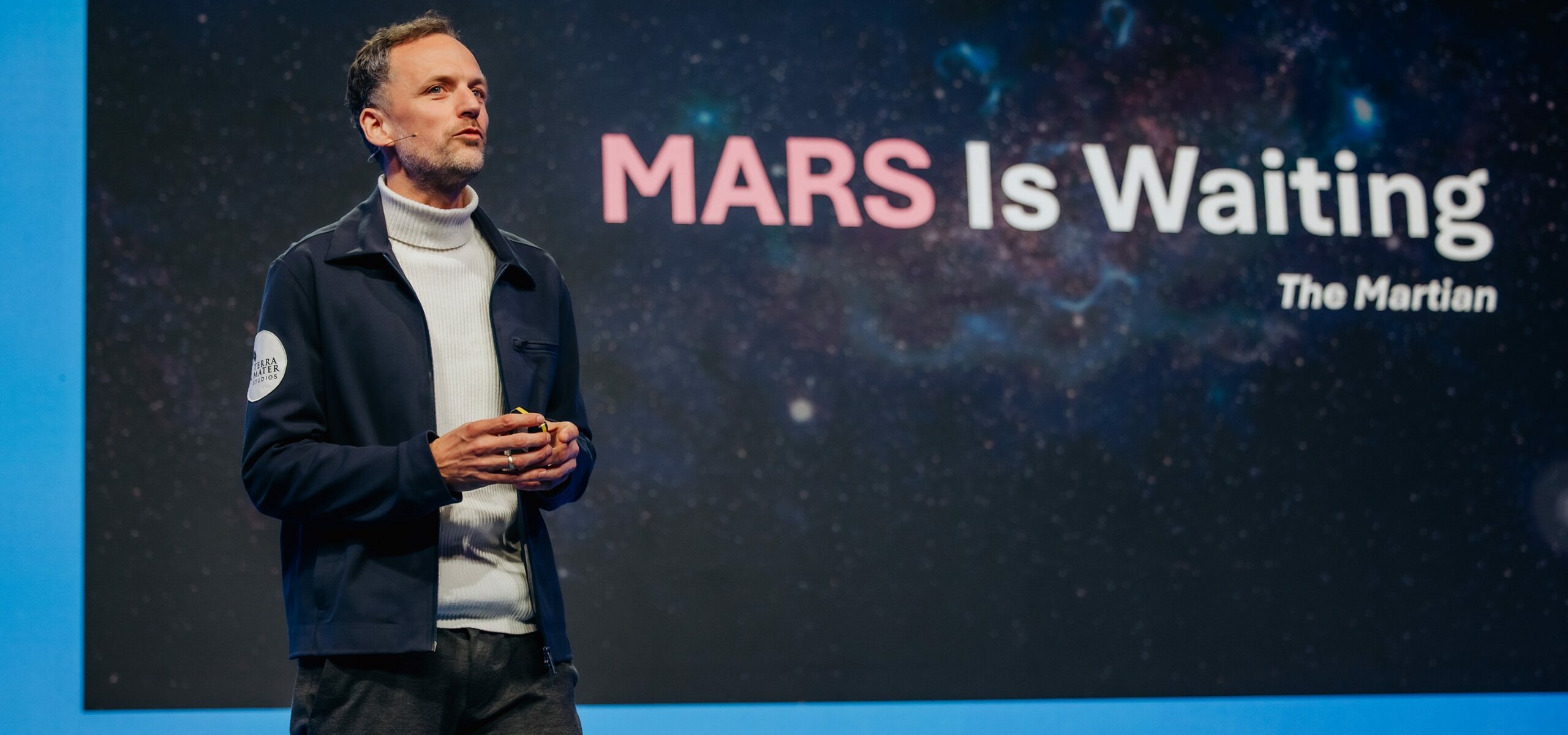 Markus Mooslechner stands on a stage in front of a large screen displaying the quote "MARS Is Waiting" from the book The Martian. Dressed in a white turtleneck and dark jacket, they hold a small device. The backdrop features a starry space image, hinting at the future of space exploration as part of the Hera Space Companion initiative.